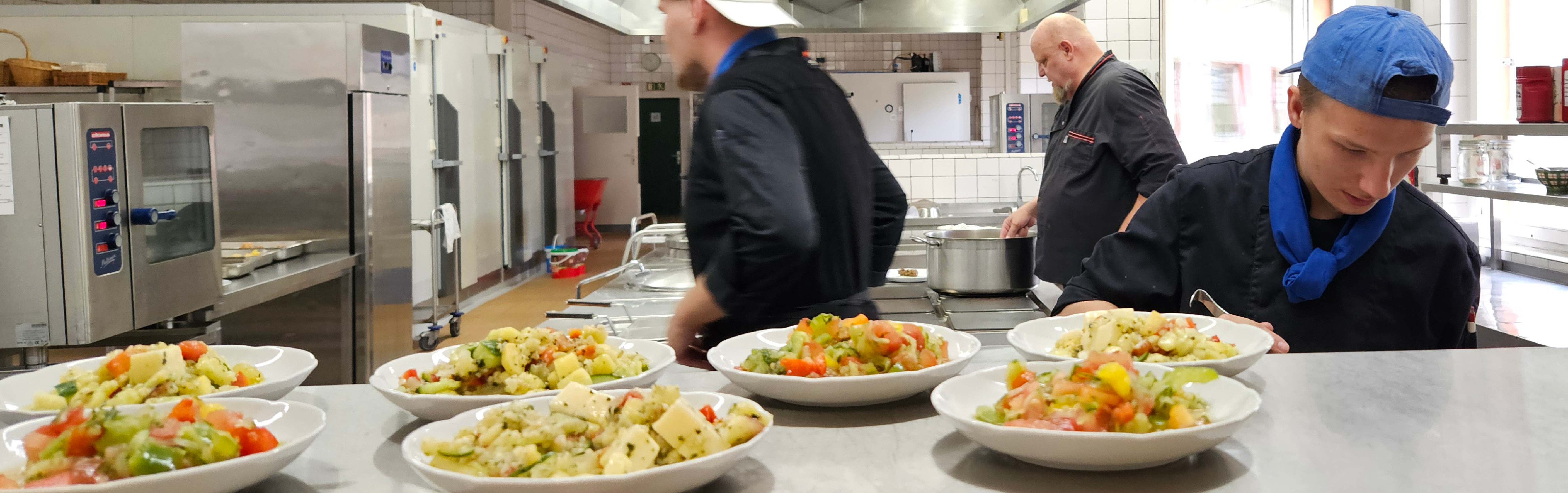 Mehrere Personen arbeiten in einer Großküche an der Zubereitung und Ausgabe von Speisen. Im Vordergrund stehen Teller mit frisch angerichteten Salaten auf einer Edelstahl-Arbeitsfläche. Im Hintergrund sind Küchengeräte, Kühlschränke und weitere Arbeitsbereiche einer professionellen Ausbildungsküche zu sehen.