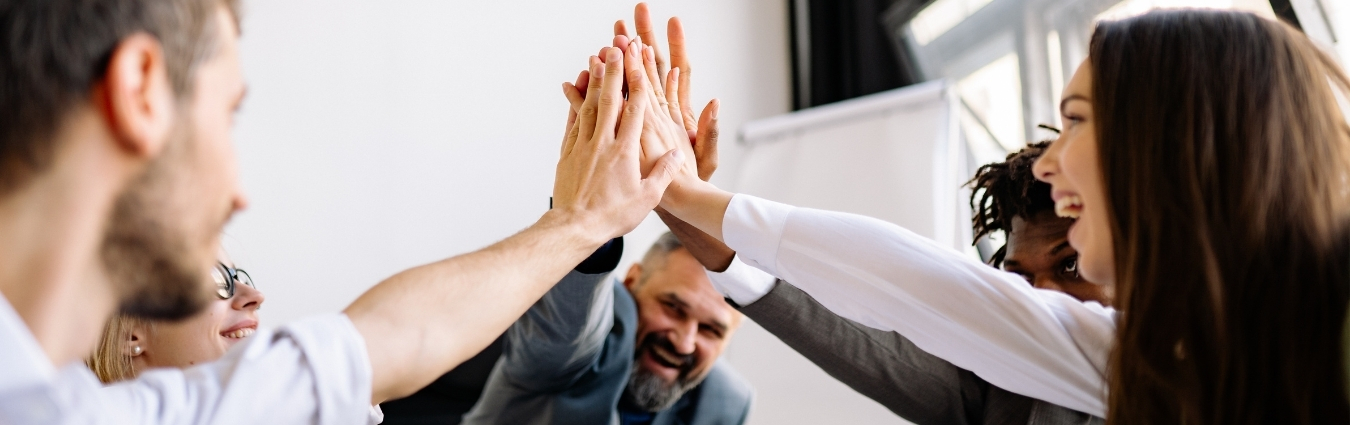 Mehrere Menschen unterschiedlicher Herkunft stehen in einem hellen Raum im Kreis und legen ihre Hände in der Mitte übereinander. Sie lächeln und wirken motiviert sowie solidarisch. Die Szene vermittelt Teamgeist, Zusammenarbeit und Gemeinschaft.