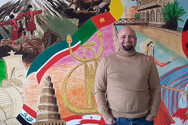 Eine Person steht in einem Raum vor einer großen, farbenfrohen Wandmalerei. Das Wandbild zeigt verschiedene Landschaften, kulturelle Symbole, historische Bauwerke und Figuren aus unterschiedlichen Ländern.