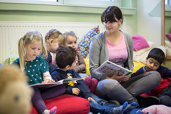 Fünf kleine Kinder sitzen zusammen mit einer jungen Frau auf Kissen auf dem Boden. Die Frau hält ein Buch in der Hand.