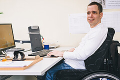 Person im Rollstuhl arbeitet an einem Schreibtisch mit Computer, Büromaterialien und Unterlagen in einem Büroarbeitsplatz.