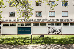 Außenansicht der Erinnerungsstätte Notaufnahmelager Marienfelde in Berlin mit Gebäudefassade, großen Fenstern und dem Schriftzug über dem Eingang, umgeben von Bäumen und einer begrünten Rasenfläche im Vordergrund.