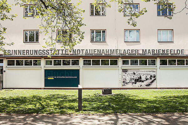 Außenansicht der Erinnerungsstätte Notaufnahmelager Marienfelde in Berlin mit Gebäudefassade, großen Fenstern und dem Schriftzug über dem Eingang, umgeben von Bäumen und einer begrünten Rasenfläche im Vordergrund.